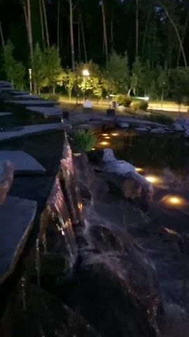 The video captures a serene nighttime scene in a park setting, likely in Barvikha, Russia. The primary subjects are a water feature with a small waterfall cascading over rocks, and the surrounding landscape. The water appears still in the pond area, reflecting the ambient light. The scene is illuminated by various light sources: warm yellow lights submerged in the pond and along its edge, and larger, spherical globe lights of white and pink hues placed on the rocky shoreline. A gazebo-like structure with a white roof is visible in the mid-ground, along with other buildings or structures further back, all softly lit.

The time of day is clearly night, with a deep blue twilight sky visible above the treeline. The weather appears calm, with no indication of wind or rain. The setting is a landscaped park with a dense forest of tall, slender trees in the background, suggesting a natural or semi-natural environment integrated with park amenities.

The overall mood conveyed is tranquil and peaceful, enhanced by the gentle sound of the waterfall and the soft, inviting glow of the lights. There are no people visible in the video. The activity is simply the presentation of a beautifully lit park at night. Notable details include the contrast between the dark water and the bright lights, and the silhouettes of the pine trees against the dusky sky. There is no visible text in the video.