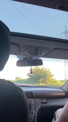This media file captures a view from inside a car driving on a rural road, likely in or around Marmarashen, Armenia. The perspective is from the back seat, looking forward through the car's interior towards the open sunroof and windshield. Sunlight streams through the sunroof, which has some water droplets or streaks on it, suggesting recent rain or mist.

The subjects are two people, whose backs are visible in the foreground. One person is wearing a dark hooded jacket, and the other, on the right, is wearing patterned shorts or a skirt. The car's dashboard and center console are also visible, showing the radio controls and air vents. A rearview mirror hangs from the ceiling, reflecting the face of the driver or front passenger. A small, red and gold dangling air freshener is suspended from the mirror.

The scene outside the car depicts a road winding through a landscape of green fields and trees, bathed in the warm, golden light of either sunrise or sunset. The weather appears to be clear with hints of moisture. The overall mood is peaceful and suggests a journey through the countryside. The visible text on the dashboard reads "SRS AIRBAG".