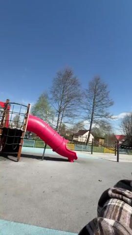 The video shows a woman standing at the base of a playground climbing structure made of ropes and metal bars. She is wearing sunglasses, a white t-shirt, and a brown plaid shirt. She is looking up at two children who are climbing on the structure. The sky is blue and sunny. There are trees and buildings visible in the background. The woman appears to be rotating the climbing structure while holding onto a metal bar.