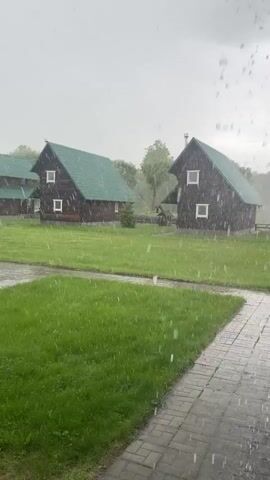 The video shows a rainy day in a rural setting. Three wooden cabins with green roofs are positioned on a green lawn. A brick walkway leads from the viewing point toward the cabins. Rain is falling heavily, obscuring the view to some extent. The overall atmosphere is grey and wet. There are no people visible.