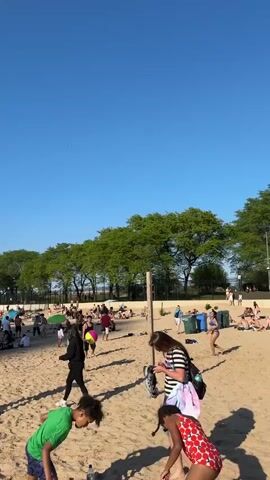 The video shows a crowded beach on a sunny day. The beach is sandy, and many people are sunbathing, walking, and playing. Some people are near the water's edge. There are boats visible in the water. Some people are playing with a beach ball. Some trees are visible lining the edge of the beach. The sky is clear and blue.