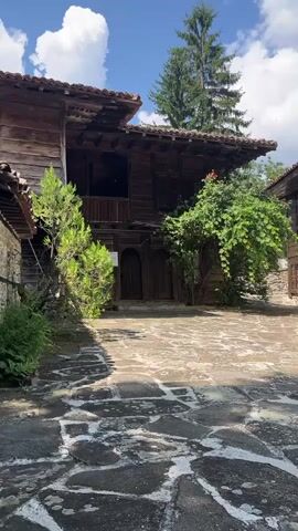 The video pans across a courtyard paved with irregular stone slabs. On the left, a multi-story wooden building with a tiled roof and balconies is visible. To the right, another wooden building with dark windows and a tiled roof is seen. Further to the right, a white building with a tiled roof and a stone archway supported by two stone pillars is prominent. The archway leads to a dark entrance. A tree with green foliage is situated in the center of the courtyard, partially obscuring the wooden buildings. The sky above is blue with white clouds. The scene appears to be an outdoor courtyard of an old architectural complex, possibly a monastery or traditional village.