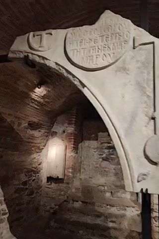 The video pans across an interior archaeological site with exposed brickwork and stone walls. A large, light-colored carved stone fragment, possibly part of an archway or lintel, is prominently displayed and supported by metal stands. The stone has inscriptions and a carved symbol resembling a pendulum. The lighting is dim, with spotlights highlighting the stone artifact and some areas of the wall.
