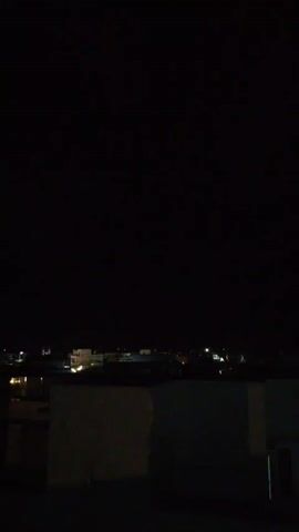 The video shows a dark night sky with a city skyline visible in the distance. There are several buildings with lights on, indicating that the city is populated. Suddenly, a flash of lightning illuminates the sky briefly.