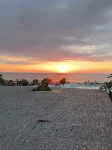 A video captures a sunset over the ocean, with a resort-like area in the foreground. The sky is filled with hues of orange, yellow, and pink, with some clouds present. The sun is setting on the horizon, casting a bright glow. In the middle ground, there's a body of water that appears to be a swimming pool, with lounge chairs and umbrellas visible around it. There are also some palm trees and other vegetation. The foreground is dominated by a thatched-roof structure, possibly a part of the resort's seating area. The scene is serene and depicts a beautiful evening by the sea.