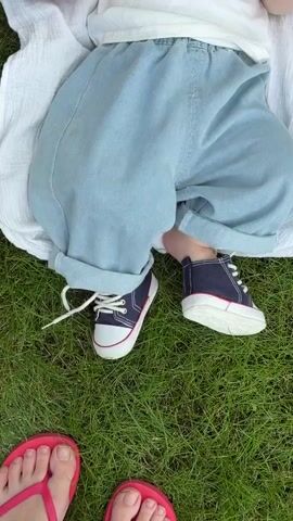 The video shows a close-up view of a baby lying on a white blanket spread on green grass. The baby is wearing a white t-shirt with the words "Good, Good Times, Times," printed in red, and light blue, baggy jeans. The baby is also wearing dark blue canvas sneakers with white laces and red trim. The camera then pans down to show two pairs of adult feet wearing pink flip-flops standing on the grass in front of the baby. The scene is outdoors on a sunny day.