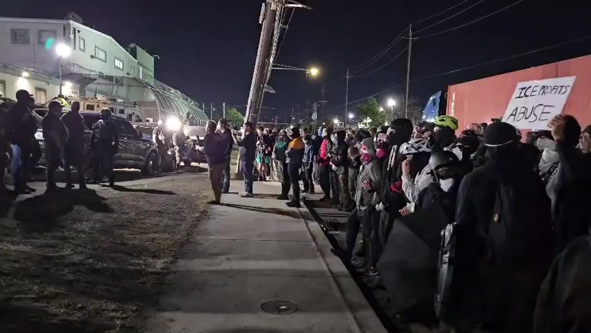 The video shows a nighttime protest.  On the left, a group of armed personnel in uniform stand on a grassy area, facing right towards a large group of protesters standing on a sidewalk. In the background, there are buildings and a vehicle with headlights. A sign held by a protester reads "ICE PROFITS ABUSE". The setting is an urban area with overhead wires and utility poles.