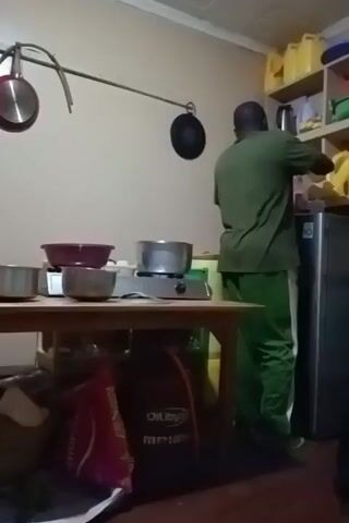 A man in a green shirt and green pants with a white stripe walks away from a refrigerator towards a table on which a gas stove, a pot, and several bowls sit. The stove is on. A bag labeled "SHOPPERS" and an "OIL Libya" gas cylinder sit underneath the table. A shelf with yellow gallon jugs sits to the right of the refrigerator. Three pans hang from a rod on the wall. The room is small.