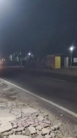 A nighttime video shows a road with a vehicle approaching and passing by. The road is dark, with streetlights visible in the distance and on the side of the road. There are trees and some structures on the side of the road, including what appears to be a yellow and red truck. The foreground of the video shows a damaged section of the road or sidewalk with broken concrete and rocks. The video captures the headlights of an approaching vehicle, which then passes through the frame, followed by a scooter. The overall scene is dimly lit and appears to be a rural or semi-urban environment.