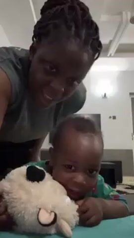A woman and a young child are in an indoor space. The child is lying on a green surface and holding a white plush toy. The woman is behind and to the left of the child. She dances while the child holds the toy and looks at the camera. The room has white walls and a ceiling with recessed lighting. A television and other furniture are visible in the background.