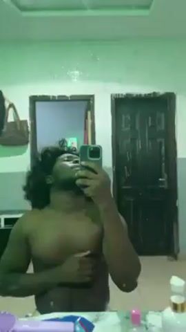 A shirtless man with a short, curly wig stands in front of a bathroom mirror, filming himself with his phone. He is dark-skinned with a slight beard and mustache. He has a muscular build. The bathroom is small, with a black door and a window visible in the mirror. The countertop in front of the mirror is cluttered with toiletries and other items, including bottles, containers, and a purple pencil case. There are sparkles on the mirror. The lighting in the room changes color slightly throughout the video, going from blue to green to red. He touches his chest and then his wig in the video.
