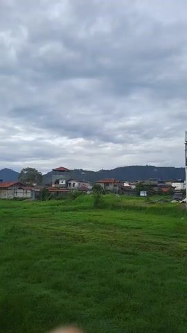 The video shows a green field in front of some houses and mountains. The sky is cloudy. The field appears to be overgrown. There are no people visible.