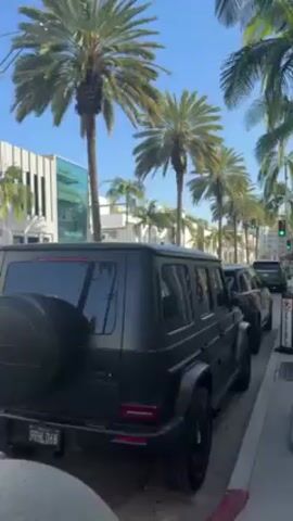 The video shows a street with parked cars and palm trees. The street appears to be in a wealthy area. A man with blonde hair and glasses on his head is holding a camera and looking at the camera. He is standing on the sidewalk next to the street. There are large, round, gray bollards lining the sidewalk.