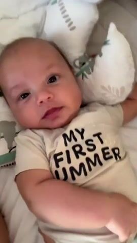 A baby with dark hair and large brown eyes lies on a white surface surrounded by patterned pillows. The baby is wearing a light-colored onesie with the text "MY FIRST SUMMER" printed on it in black lettering. A person's hand with pink nail polish reaches into the frame and gently touches the baby's arm. The baby's expression shifts from neutral to a slight smile and then a wider grin. The camera angle is overhead, looking down at the baby.