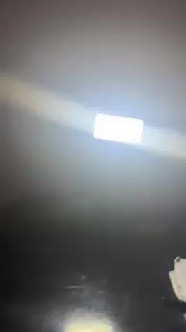 The camera points upwards at a bright rectangular light fixture set into a dark ceiling.  The light casts a strong, radiant glow downwards.  To the bottom right of the frame is a white object, possibly paper or cloth.  A small portion of wood trim is visible in the upper left corner.  The background is mostly a dark grey gradient.