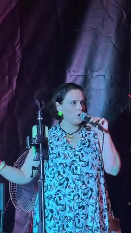 A woman stands on a stage in front of a dark, wrinkled backdrop, holding a microphone and speaking. She has short, dark hair and is wearing a sleeveless dress with a black, white, and red abstract pattern. She also has bright green earrings and a necklace with an eye-shaped pendant. Her left hand is raised, and her right hand holds the microphone close to her mouth. To her left, a microphone stand is visible, along with a cell phone attached to it and a portion of a fan below. The lighting on the stage is dim with areas of pink and green light.