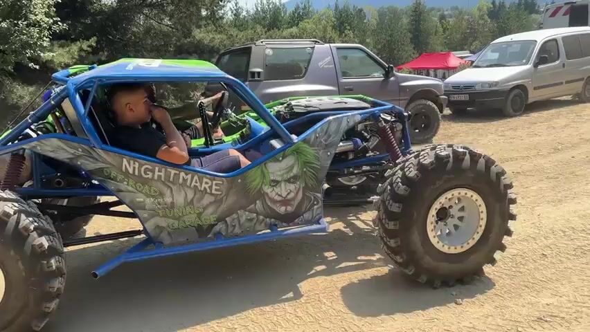 A blue and green off-road buggy with large tires sits on a dirt road. The buggy has a roll cage and the side features graphics of the Joker and the words "NIGHTMARE" and "OFFROAD TUNING". A man is sitting in the driver's seat. In the background, there are other vehicles, including an SUV, a van, and an ambulance. There are also trees and mountains in the distance. The scene appears to be an outdoor event or gathering.