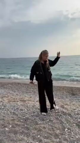 A young woman in black pants and a black jacket twirls happily on a rocky beach, the sea and a cloudy sky behind her. The scene then shifts to a wider view of the beach, revealing mountains in the background and a few people scattered along the shore. Finally, a woman with dark hair and a grey jacket smiles directly into the camera, standing on the same rocky beach.