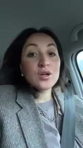 The video shows a woman in a car, wearing a gray blazer and a silver chain necklace. She is also wearing a seatbelt. She makes exaggerated lip pursing gestures, then makes a grimace. The setting appears to be inside a car during the day, with a glimpse of a rainy window.