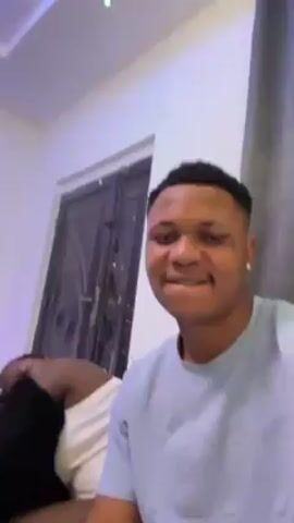 The video shows a man recording a selfie video with another person asleep in the background. The man in the foreground is wearing a light blue t-shirt and has short, dark hair. He is smiling and looking directly at the camera. He appears to be indoors, possibly in a living room, as there is a door and a framed picture visible on the wall. Behind him, another person is lying on a grey couch, covered with a black fuzzy blanket. The room has white walls and a light-colored ceiling.