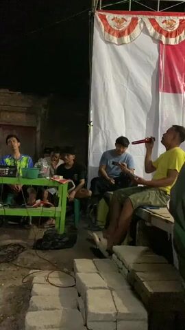 A group of young men are gathered outdoors at night, in what appears to be a casual setting. In the foreground, a man in a yellow t-shirt is seated on a wooden bench, holding a microphone to his mouth and speaking or singing. He is wearing patterned shorts and flip-flops. To his left, another young man in a blue t-shirt is seated, looking at his phone. Further to the left, a group of men are seated around a green table. One man, wearing a blue and yellow jacket, is seated behind what appears to be sound equipment. Another man in a black t-shirt is seated to his right. The background shows a white banner with red and orange trim, possibly decorated with a pattern. The ground is uneven, with some concrete blocks visible in the foreground. The overall lighting suggests it is nighttime, with some artificial illumination present.