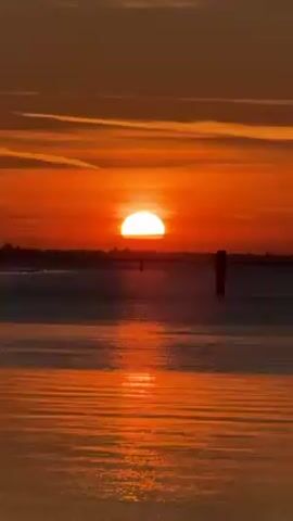The video shows a vibrant orange sunset over a body of water. The sun is low on the horizon, appearing as a semi-circle due to its partial submersion. The sky above is a mix of orange and yellow hues, with streaks of clouds. The water reflects the colors of the sunset, creating a shimmering orange surface. There are a few wooden posts or markers visible in the water.