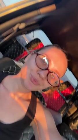 A woman with her hair in a bun and wearing a black tank top and round glasses takes a selfie in the back of a car. The car has a sunroof and a decorative, diamond-patterned floor mat. The back of the car also appears to be packed with items, including red and white objects. A child in a red shirt is briefly visible behind the woman. Sunlight illuminates the woman's face, giving it a warm glow. Throughout the video, she poses and smiles slightly.