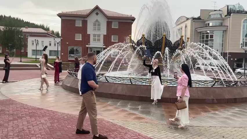 The video captures an outdoor scene near a large, ornate fountain in a public square. The fountain is the focal point, with water spraying upwards in a dramatic display. Several individuals, mostly women dressed in formal attire, are positioned around the fountain. On the left side of the fountain, a group of women in long dresses are posing and interacting. One woman in a dark dress stands near the edge of the frame, while others in lighter colored dresses are further back. A man in a blue shirt and tan pants stands facing the fountain, his back to the camera, observing the scene. To the right of the fountain, two women are interacting, one in a pink top and white pants, the other in a black top and white skirt. In the background, buildings with red and white facades are visible, along with trees on a hillside. The ground is paved with red and pinkish-brown bricks in a patterned arrangement. The overall atmosphere appears to be a gathering or event, possibly a celebration or photo opportunity.