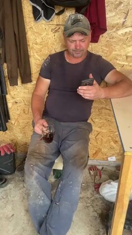 A man sits on a stool in a room with plywood walls, holding a phone in his right hand and a glass of dark liquid in his left. He wears a dark t-shirt, a baseball cap, and dirty blue work pants. Several items hang on the wall behind him, including a dark jacket and a red cloth. Various objects, including work gloves, are scattered on the floor. He appears to be looking at his phone.