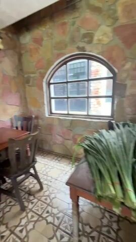 The video shows an outdoor seating area of a restaurant. The walls are made of rough-cut stones in various shades of pink, yellow, and brown. An arched window with multiple panes looks out onto a brick building. The floor is tiled with a geometric pattern. Several wooden tables and chairs are arranged throughout the space. In the foreground, a rectangular wooden table is set with four dark wooden chairs. To the left, a woman in a white top and plaid shorts is seated at a table, looking at her phone. Behind her, a covered patio area with exposed beams and roof tiles is visible, with lush greenery and trees beyond. A ceiling fan is mounted on the patio roof. The lighting is natural, suggesting daytime.