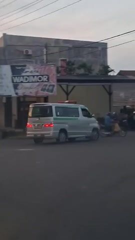 The video shows a silver ambulance with green and red markings driving on a road. A motorcycle with a rider is seen near the ambulance. In the background, there are buildings, one with a sign that says "DIMOR". The sky is overcast, suggesting it might be evening or early morning. The ambulance has its lights on, and the overall impression is of a vehicle in motion.