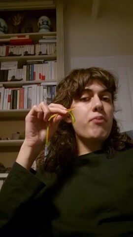 A medium shot depicts a woman with brown curly hair, wearing a black top, positioned against the backdrop of a bookshelf. She is in the process of eating a yellow and green gummy candy. The bookshelf, painted white, features various decorative items and books arranged on its shelves. The woman’s face is the primary focus as she chews the candy, maintaining eye contact with the camera.