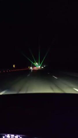 The video depicts a nighttime drive on a highway. The perspective is from inside a car, looking through the windshield. The road is paved, with white lane markings visible. On the left side of the road, there's a line of orange traffic cones and a yellow sign. In the distance, several cars are visible, their headlights illuminating the road.

The most striking feature of the scene is a set of bright green laser lights projected upwards into the night sky from a point ahead on the highway. These lasers create a fan-like effect, emanating from a central point and spreading out. In some frames, the lasers also exhibit red and blue hues, suggesting a multi-colored light display.

The time of day is clearly night, with the sky being completely dark. There are no visible weather conditions like rain or fog. The setting appears to be a highway outside of a populated area, given the darkness and the presence of only a few cars. The overall mood is somewhat futuristic or festive due to the unusual laser lights.

The visible text at the bottom of the screen is part of the car's dashboard, displaying speed readings in kilometers per hour, with numbers such as "120", "140" visible. The location context provided is Zarechnoye, Kazakhstan.