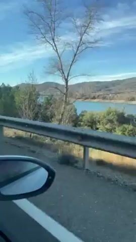 The video appears to be taken from inside a moving vehicle, showing the roadside scenery. A guardrail is visible in the foreground, partially obstructing the view. Beyond the guardrail, there is a mix of dry grass and trees with varying degrees of foliage. In the distance, a body of water is visible, possibly a lake or reservoir. Beyond the water, there are hills or small mountains under a blue sky with scattered clouds.