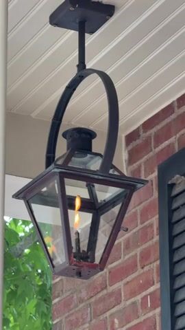 A video shows a close-up shot of a gas lantern hanging from the porch ceiling of a brick house. The lantern has a dark metal frame with glass panes and a lit flame inside. A green leafy tree is visible on the left side of the frame, and part of a black shutter is visible on the right. The ceiling appears to be made of white wooden planks. The lantern swings gently as the video progresses.