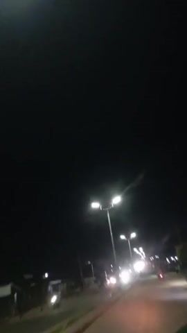 The video shows a night scene from the perspective of a moving vehicle, likely a motorcycle, on a paved road. Streetlights illuminate the road, casting bright white light. Other vehicles, including cars and motorcycles, are visible on the road, their headlights and taillights creating streaks of light due to the motion. Some buildings or structures can be seen along the side of the road. The sky is dark, indicating it is nighttime. The overall impression is of a typical urban or suburban environment at night with ongoing traffic.