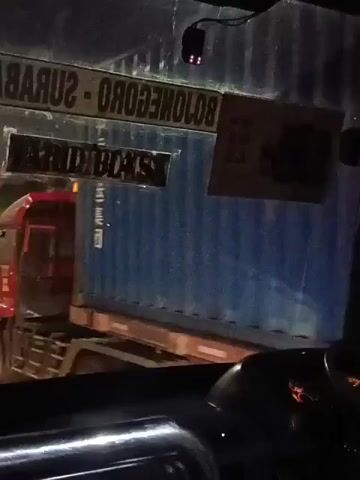 The video begins inside a vehicle, looking out the windshield. A red truck with a blue shipping container is visible in the distance, passing by. The truck is illuminated by its headlights and some ambient light. As the video progresses, the perspective shifts slightly. The view changes to looking out a side window of the same vehicle. A man wearing a cap and a patterned shirt is sitting in the foreground, facing away from the camera. Outside the window, a tanker truck with red and white markings is seen driving on a highway at night. The tanker truck has its taillights and emergency lights illuminated, indicating it might be a fire truck or a similar emergency vehicle. The surrounding environment appears to be a dark, hilly landscape with streetlights and road signs visible along the highway. The video is shaky and seems to be filmed by someone in motion.