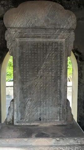 The camera begins very close to a stone tablet with Asian script, gradually pulling back to reveal an enclosed stone pavilion with decorative archways. The pavilion is decorated with intricate carvings and designs, and glimpses of greenery can be seen through the archways, suggesting an outdoor setting.