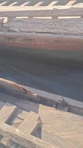 The video shows a man sanding the inside of a wooden boat. The boat is resting on a wooden scaffolding-like structure. The man is wearing orange shorts and sanding with a small electric sander. The boat appears old and the wood is weathered. In the background, there is a sandy area with some green vegetation. The video seems to be taken during a sunny day.