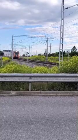A train with orange and black markings approaches on a track under a cloudy sky. The train is seen from a road, with a metal guardrail and grass in the foreground. There are power lines and poles visible above the tracks. The train passes by and continues down the track.