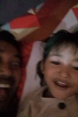 A man and a young child are lying down. The man is on the left, his face close to the camera with his mouth open and eyes wide. The child is on the right, also smiling broadly and looking towards the camera. They are both on a bed with a patterned red and white sheet. The child is wearing a light-colored top. The man is wearing a dark shirt. In the latter part of the clip, the man is holding a smartphone, which appears to be displaying a game, and the child's arm is resting over his. The lighting is dim.