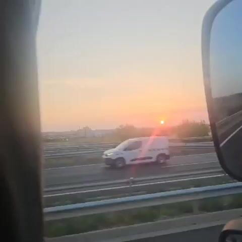 The video shows a sunset viewed from a moving vehicle. The sun is a bright, orange orb low in the sky, casting a warm glow and a prominent lens flare across the frame. In the foreground, a portion of the vehicle's interior is visible on the left, and a side mirror is on the right, reflecting the road and sky. The landscape outside consists of a highway with faint lane markings, bordered by a metal guardrail and some vegetation. In the middle ground, there are fields and scattered trees, with some buildings and a distant hill line. The sky is a gradient of soft orange and pink.