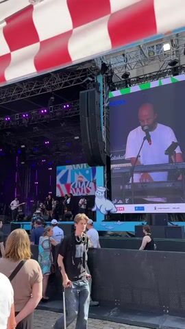 The video shows an outdoor music festival, likely in Kiel, Germany, judging by the visible text "KIEL" on a screen and banner. The scene is set under a red and white checkered canopy. A band is performing on a brightly lit stage, with various instruments and stage lighting visible. On a large screen to the right, a man is shown close-up, playing a keyboard and singing into a microphone. He is wearing glasses and a white t-shirt. Several people are gathered in front of the stage, watching the performance. Some are leaning against a barrier, while others are standing. The weather appears to be sunny, and the time of day is likely afternoon or early evening, given the bright lighting. The mood seems to be relaxed and engaged, with people enjoying the music. Visible text includes "KIEL," "WINS," "MARSHALL," "REWE," and "WELLER." A cartoon seal graphic is also prominently displayed on a blue banner in front of the stage. A young man with headphones, using crutches, is walking in the foreground, also observing the event.