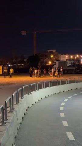 A group of people are gathered on a paved area at night. A crane is visible in the background, along with buildings and lights. There are also vehicles present, including a large truck. The ground is marked with white dashed lines, suggesting a road or pathway. Metal railings line the edge of the paved area.