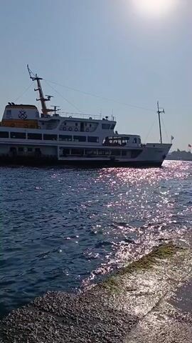 A large white passenger ferry with multiple decks moves across blue water under a clear sky. The sun reflects brightly on the water's surface. The ferry has life preservers visible along its upper decks. In the foreground, a textured concrete or stone embankment is visible on the right side of the frame. The ferry appears to be moving from left to right.