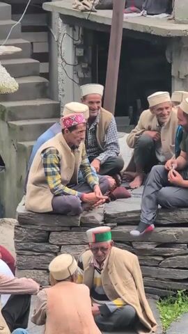 A group of men are gathered outside, sitting on stone steps. They are all wearing traditional caps, and some have on vests. They appear to be in conversation, with some smiling and looking at each other. The environment seems to be a rural or mountainous village, with stone buildings and stairs visible in the background. The lighting suggests it is daytime, possibly overcast. The camera is positioned at a medium distance, capturing the scene from a slightly elevated angle.
