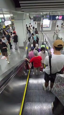 A medium shot captures a busy subway station with people on an escalator. The escalator's metallic steps and yellow safety strip are prominent in the foreground. A woman in a red shirt and grey pants, carrying two shopping bags, is ascending the escalator. Behind her, a person wearing a white t-shirt and dark shorts, with a tan baseball cap and a backpack, is also on the escalator, looking at their phone.

In the background, other passengers are visible on the escalator and on the station platform. Some are walking, while others appear to be waiting. The station is brightly lit with fluorescent lights on the ceiling. Signage is visible on the walls, including directional arrows with Chinese characters and a digital display screen showing text. One sign has an arrow pointing left with Chinese characters, and another sign has an arrow pointing right with Chinese characters and "往山阳路" which translates to "Towards Shanyang Road."

The overall scene suggests a typical day in a public transportation hub, likely during a time of moderate foot traffic. The atmosphere appears orderly, with people moving purposefully. The time of day is not explicitly indicated, but the bright, artificial lighting suggests it could be anytime. The weather is not discernible from the indoor scene. The setting is clearly a modern subway station in Gongshu District, China, evidenced by the language on the signs and the architectural style.