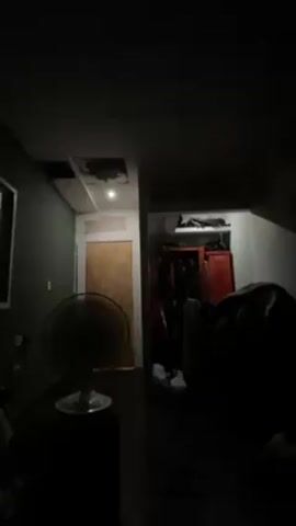 The video shows a dark room with some light sources. There is a ceiling fan in the foreground and a doorway in the background, through which one can see a closet. There is a hole in the ceiling with wires and pipes visible. The room looks somewhat messy, with items scattered in the closet and on the floor. The lighting is poor, making it difficult to see details.