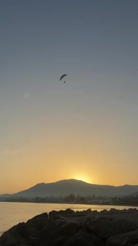 A paramotorist is seen in flight against a dusky sky. The sun is setting behind a silhouetted mountain range, casting a warm orange glow across the horizon. In the foreground, a rocky shoreline borders a body of water. The paramotorist, a small figure suspended beneath a colorful parachute, is the sole focus of attention in the vast expanse of the sky. The scene captures a moment of serene solitude and aerial adventure as daylight fades.