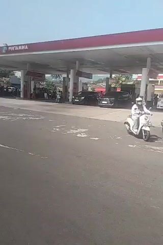 The video shows a gas station with several cars and motorcycles. A white scooter is being ridden by a person wearing a white helmet and a white shirt. Another motorcycle with two people, one wearing a black helmet, is passing by. The gas station is branded as PERTAMINA. The sky is clear and the weather appears to be sunny. The road in front of the gas station is paved and shows signs of wear. There are also some trees and buildings visible in the background.